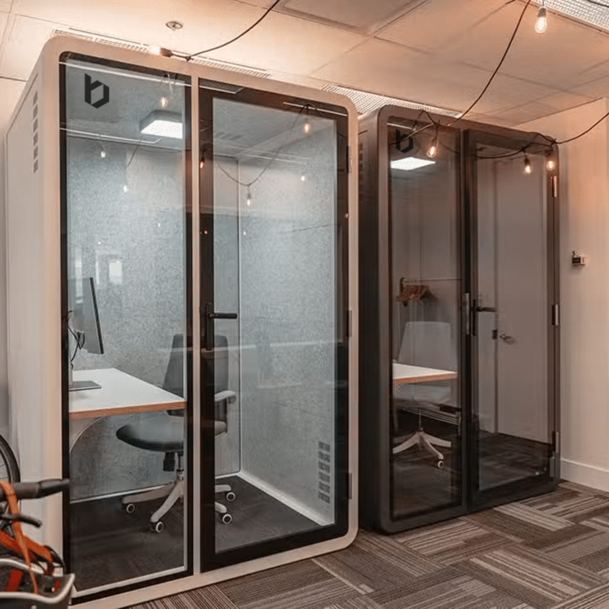 Two modern office acoustic pod cabins with glass doors and white/light gray exterior frames, shown installed in an office interior with desks, chairs, carpet, and hanging string lights visible.