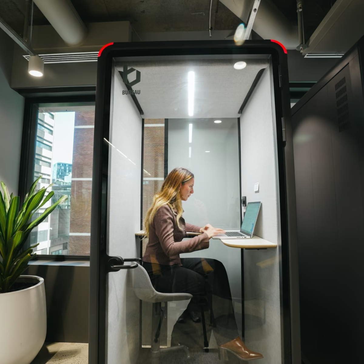 A woman sits inside a glass privacy call room with a built-in desk and chair, working on a laptop. The room has a black frame, clear glass walls, and a small window on the left showing an office setting with a plant.