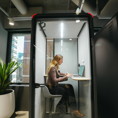 A woman sits inside a glass privacy call room with a built-in desk and chair, working on a laptop. The room has a black frame, clear glass walls, and a small window on the left showing an office setting with a plant.