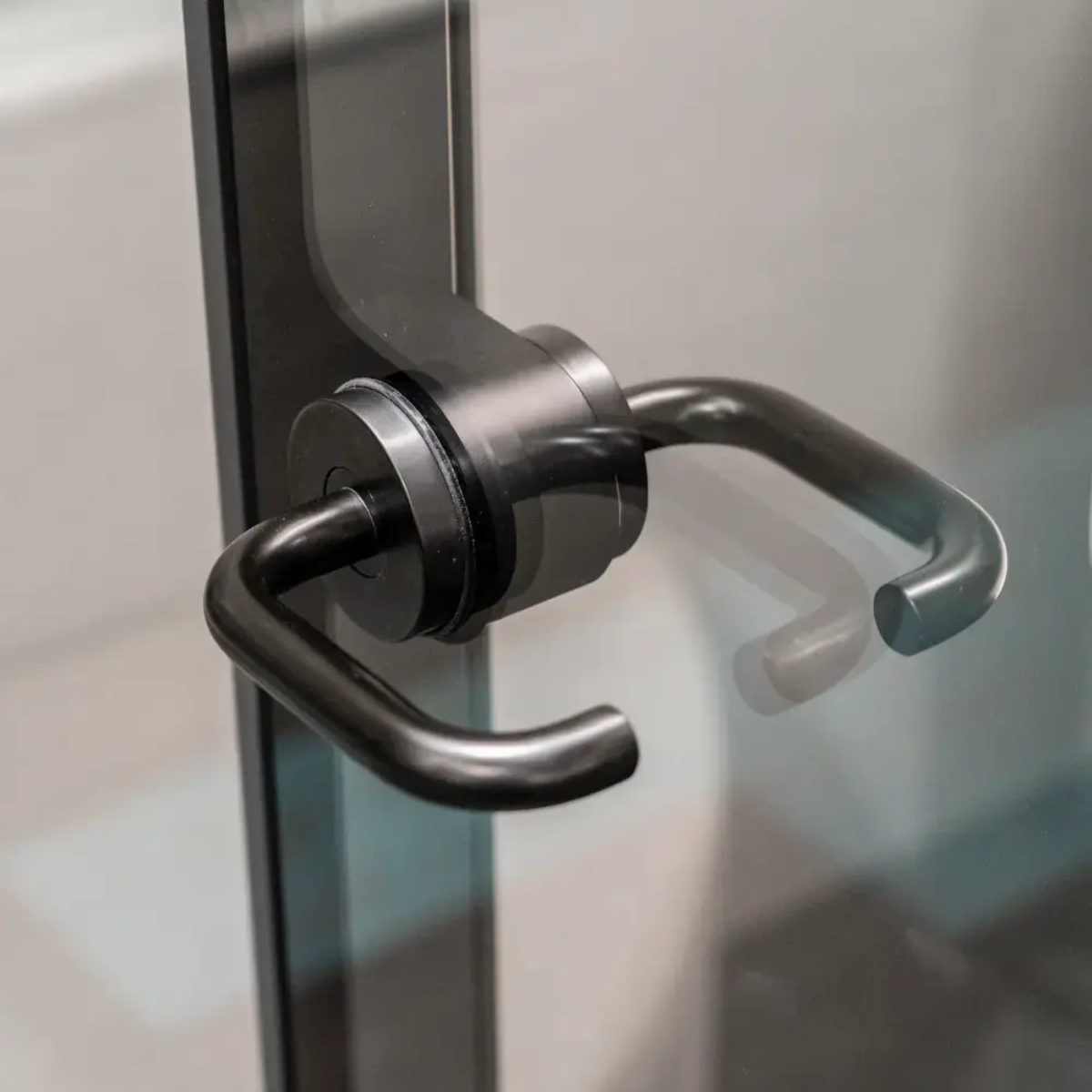 A close-up of a matte black metal door handle with a cylindrical mounting base, attached to a glass door frame.