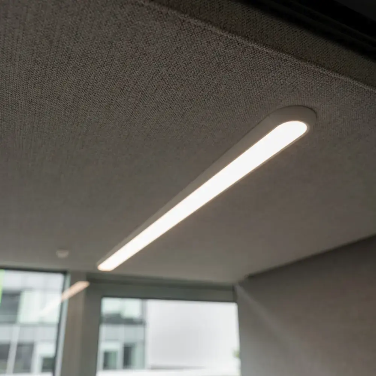A modern linear LED ceiling light fixture installed in a textured acoustic ceiling panel, shown illuminated in an office or interior space.