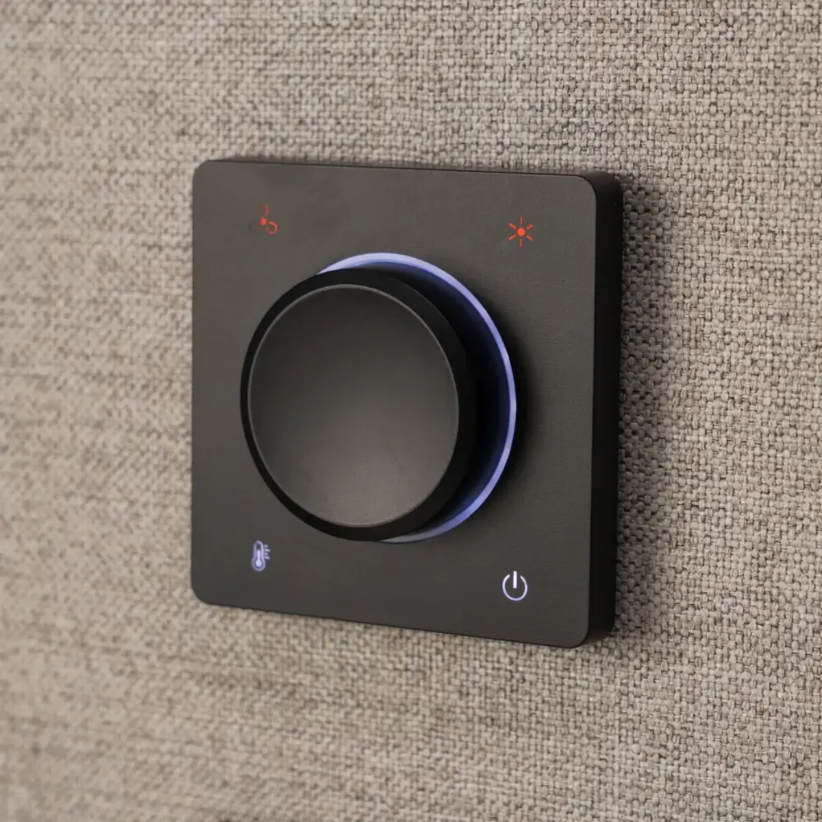 A square black smart home control panel with a central rotary knob, mounted on a textured beige fabric surface. The panel features illuminated icons for brightness, temperature, and power.