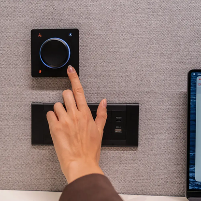 A hand pressing a touch control panel mounted on a textured gray wall, with a second panel below it and part of a smartphone visible at the right edge.