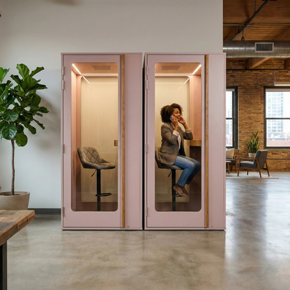 Person using a pink phone booth in an office setting with a plant and desk in the background.